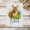 Charming watercolor birthday card showcasing an Abyssinian cat in a natural scene, complete with a "Happy Birthday" note.