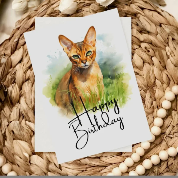 Delightful watercolor card featuring an Abyssinian cat in a natural setting, celebrating with a "Happy Birthday" message.
