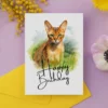 Printable birthday card with a watercolor illustration of an Abyssinian cat in nature, adorned with a "Happy Birthday" greeting.