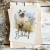 Delightful watercolor illustration of a Birman cat in a natural scene, designed as a birthday card with "Happy Birthday" text.