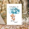 Charming card showing a flying teddy bear with blue balloons, featuring the text, "Welcome to the world, little one!"