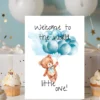 Cute teddy bear holding blue balloons on a card that says, "Welcome to the world, little one!" in watercolor style.