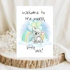 Watercolor baby shower card featuring a koala on a cloud with balloons and a rainbow, saying "Welcome to the world, little one!"