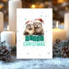 Printable Christmas card with two otters in Santa hats and a snowy winter background