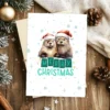 Digital Christmas card with two smiling otters in Santa hats and snow-covered background