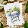 Watercolor illustration of a swan on a birthday card, surrounded by nature, featuring a festive "Happy Birthday" message.