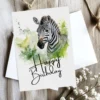 Cheerful zebra birthday card illustrated in watercolor, set outdoors, with a bright "Happy Birthday" text.